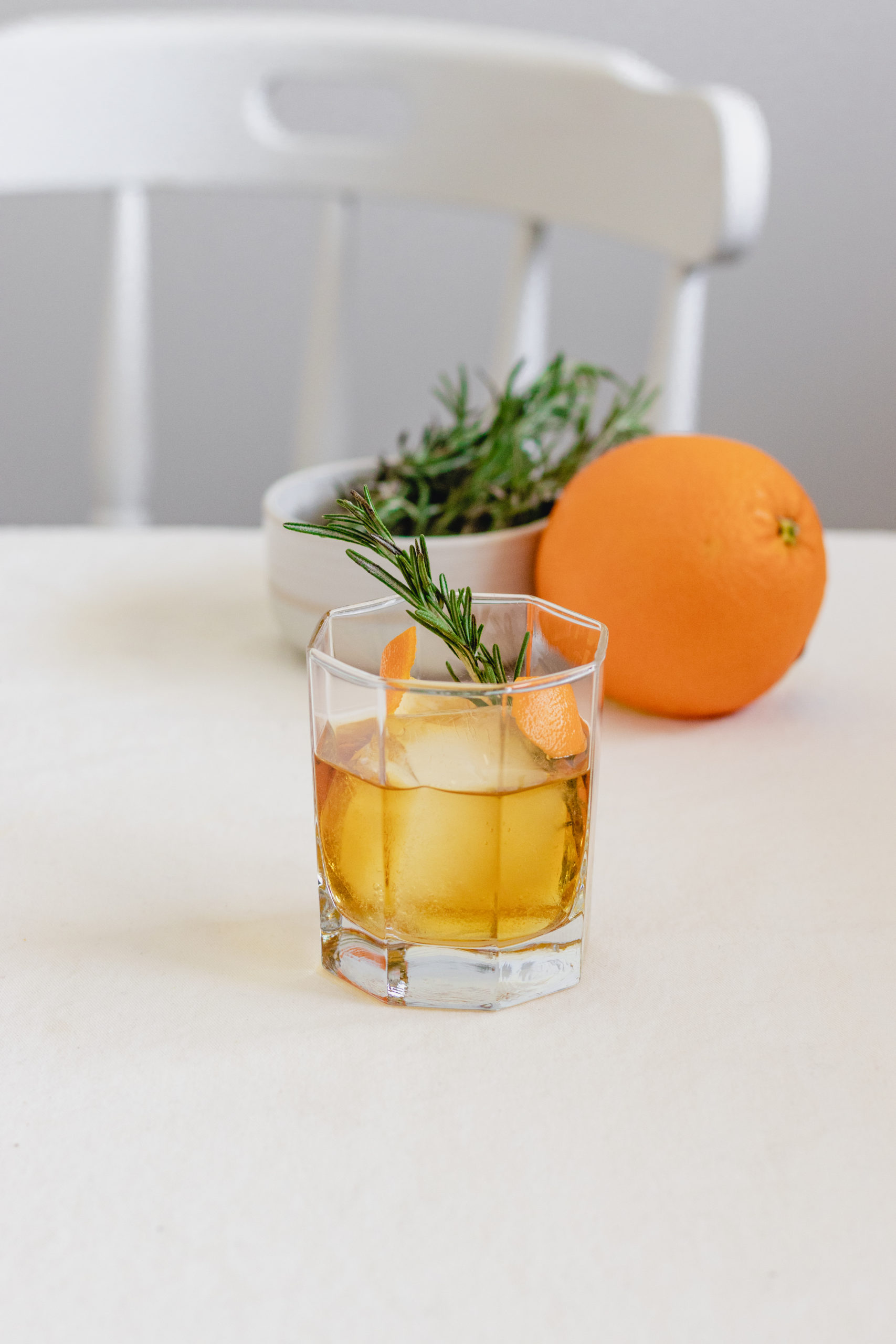 Whiskey Cocktail on a white table with an orange and fresh rosemary in the background